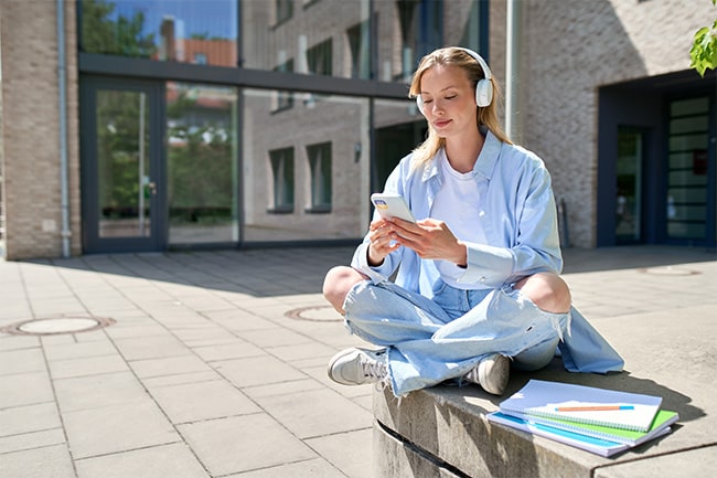 Student med hörlurar sitter på stenmur och tittar på mobil.
